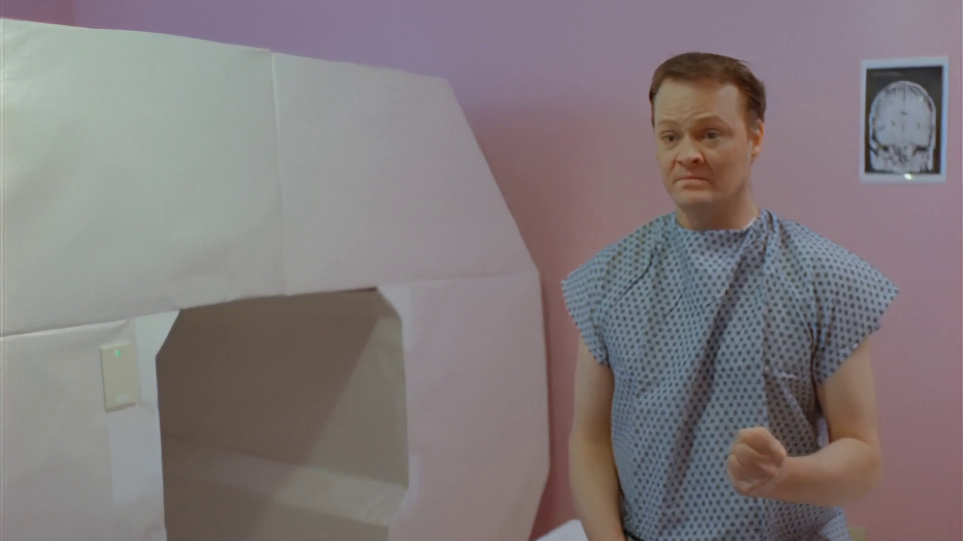 Color photo, a screenshot from the film After Last Season. In it a man sits in a pink room looking either concerned or confused. He is dressed in a hospital gown and his left hand is half-raised in a closed fist. He sits on a table next to a large prop MRI machine obviously made of white paper and cardboard.