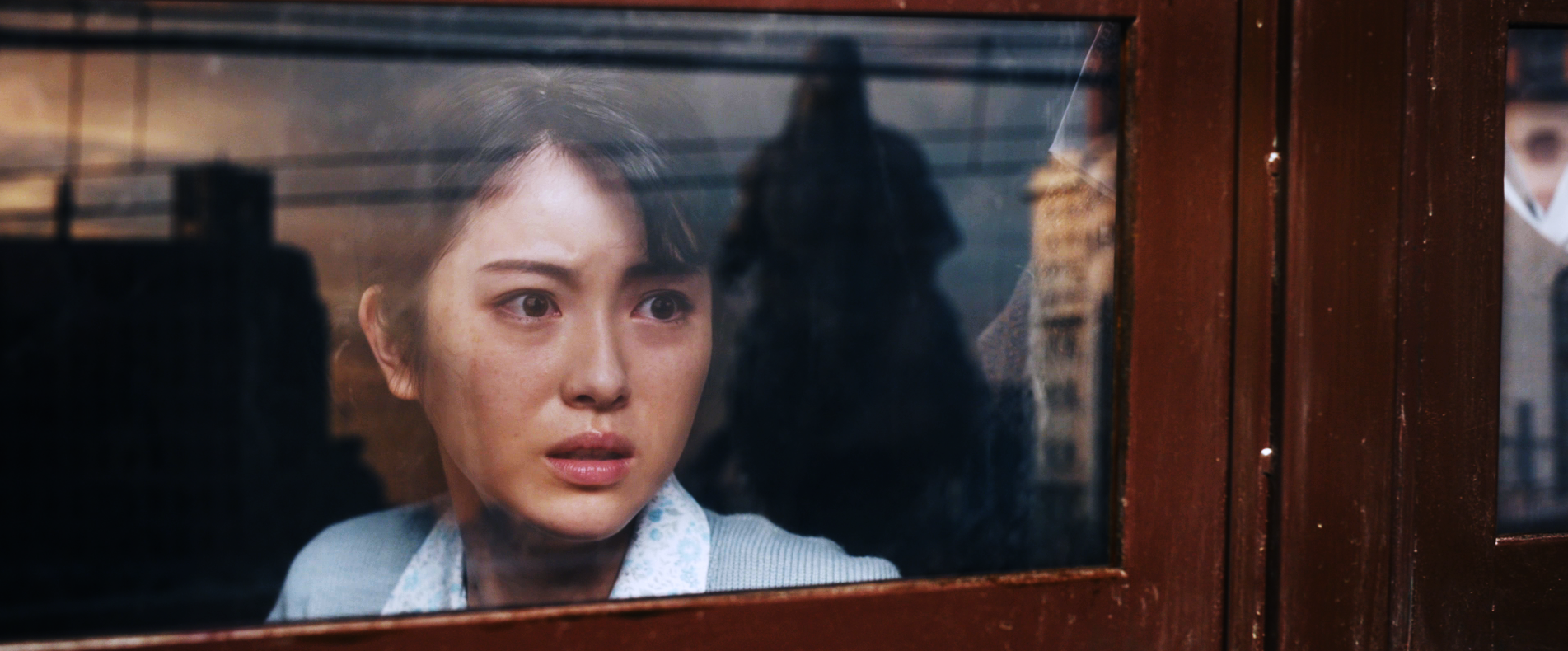Color photo, a screenshot from the film Godzilla Minus One. A woman looks out of the window of a train car, frightened. In the reflection of the window is the shadowy form of the approaching Godzilla.